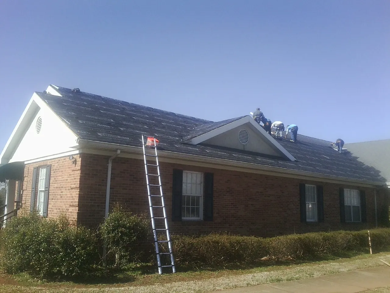Crew tearing shingles off a brick ranch roof with a ladder set — Metal Roof Repair work by our crew in Hills