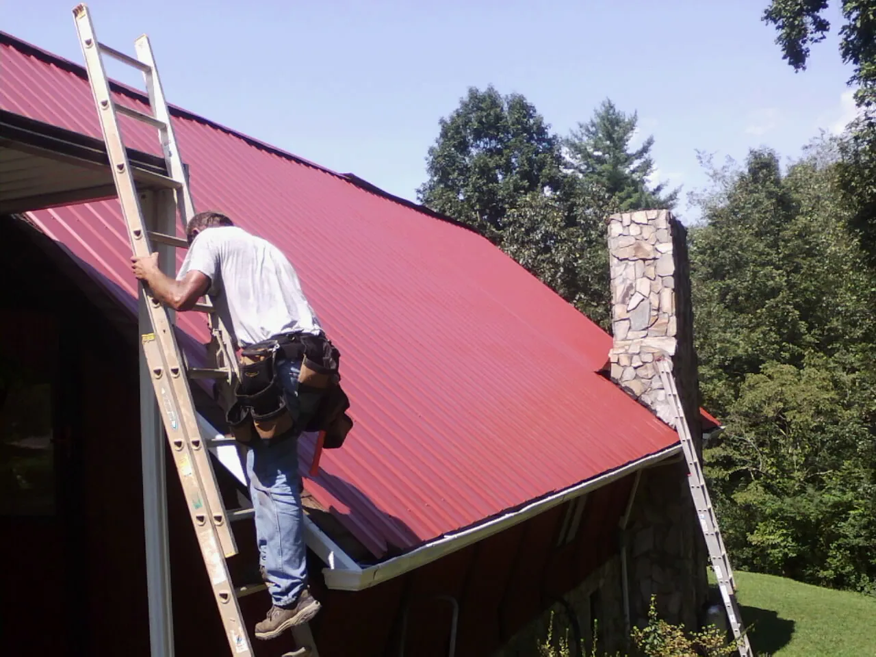 Roofer on a ladder beside a red metal roof and stone chimney — Soffit & Fascia Repair by Roofing Brothers in Hills